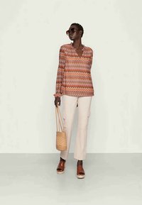 Blusa de mangas longas com padrão em chevron laranja e rosa, combinada com calças cargo claras, sandálias de cunha castanhas e uma bolsa de palha.