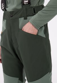 Donkergroene cargobroek met lichter groene accenten, voorzien van zijzakken en verstelbare banden, gemaakt van duurzaam, flexibel materiaal.