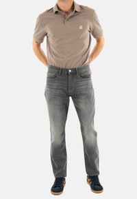 Camisa polo beige con logo en el pecho, combinada con jeans de mezclilla gris. El calzado presenta un diseño azul oscuro con acentos en marrón claro.