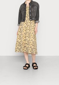 Černá denimová bunda přes žluté, vzorované midi šaty s záhyby. Černé sandály s detaily přezky. Neutrální pozadí.