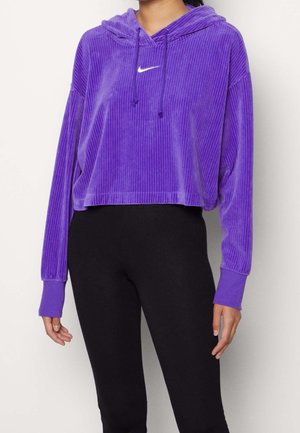 Personne portant un sweat à capuche Nike violet côtelé avec un logo swoosh blanc et des leggings noirs ajustés sur un fond uni.