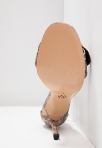 Chaussure à talons hauts avec motif en peau de serpent, vue de la semelle, appuyée contre un coin de mur et de sol blanc.