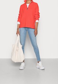 Korallfärgad zip-neck tröja med ribbad struktur, vit långärmad skjorta under, kombinerad med ljusblå skinny jeans och vita sneakers, bär en beige shoppingväska.