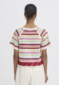 Žena s vlasmi zviazanými do drdolu, oblečená v farebnom pruhovanom háčkovanom top s krátkym rukávom a krémových nohaviciach, zobrazená zozadu.