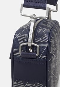 Sötétkék szintetikus táska geometrikus mintákkal, ezüst színű fém kiegészítőkkel és textilezött pánttal. Cipzáros záródással és textúrált felülettel rendelkezik.