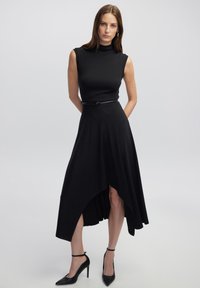 Schwarzes ärmelloses Kleid mit hohem Kragen, asymmetrischem Saum und seitlichem Schlitz. Es hat ein tailliertes Oberteil und wird mit einem dünnen Gürtel betont. Kombiniert mit High Heels.