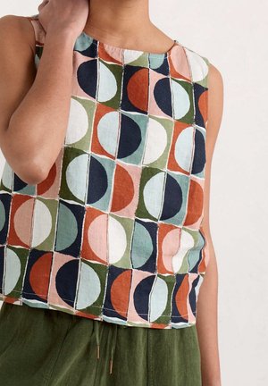 Ärmelloses Oberteil mit geometrischem Muster aus Kreisen und Quadraten in Marineblau, Rost, Grün, Weiß und Pink, getragen zu olivgrüner Hose.