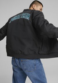Sort bomberjakke med "PUMA TEAM" brodert i teal på baksiden, med en glatt tekstur og ribbestrikket kant. Pairt med blå jeans.