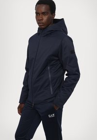 Giacca a hood blu navy con texture liscia, tasche laterali con zip e un logo sulla manica. Indossata con pantaloni sportivi coordinati.