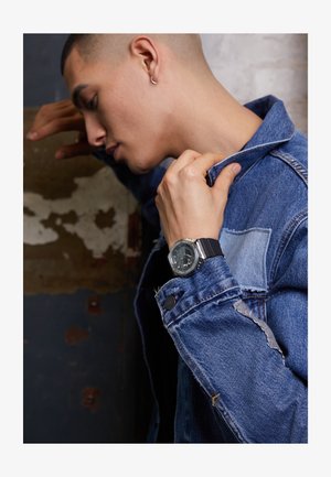 Schwarze analoge Uhr mit rundem Zifferblatt am Handgelenk, kombiniert mit einer blauen Jeansjacke mit fransigen Details und einer aufgesetzten Tasche.