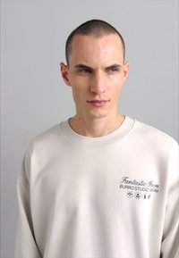 Kremfarget sweatshirt med ribbestrikket hals. Har tekstdetaljer foran i mørkegrønn. Glatt tekstur, avslappet passform, minimalistisk design.