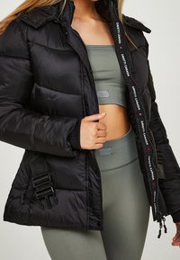 Chaqueta de plumón negra con un acabado brillante, que cuenta con un cierre frontal con cremallera, capucha y logo bordado. Incluye correas ajustables en los laterales.