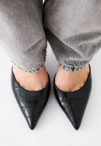 Zapatos negros de punta afilada con una textura brillante embosada de cocodrilo, combinados con pantalones de mezclilla gris, remangados.