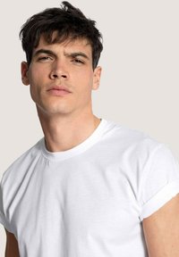 Weißes Baumwoll-T-Shirt mit kurzen, umgeschlagenen Ärmeln, Rundhalsausschnitt und glatter Textur. Das Gesicht des Models ist leicht zur Seite geneigt und zeigt einen neutralen Ausdruck.