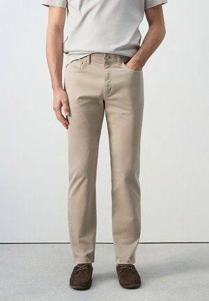 Hombre con pantalones beige y zapatos marrones de ante con cordones, de pie con una mano en el bolsillo frente a un fondo liso.