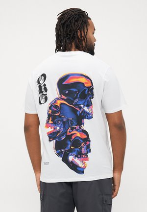Weißes T-Shirt mit kurzen Ärmeln, das auf der Rückseite eine lebendige Grafik von überlappenden Schädeln in Blau- und Orangetönen zeigt, mit Text an der Seite.