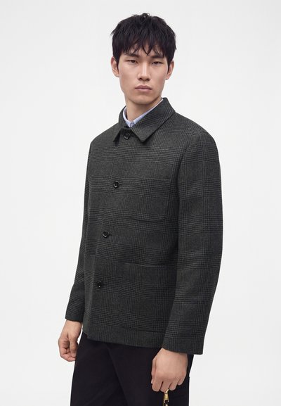 Homme portant une veste boutonnée à carreaux gris foncé avec col, debout devant un fond clair uni.