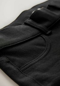 Schwarzer Stoff mit einer glatten Textur, ausgestattet mit einer geschwungenen Tasche und gestickten Details. Ein Verschlussband ist sichtbar und befindet sich am oberen Rand.