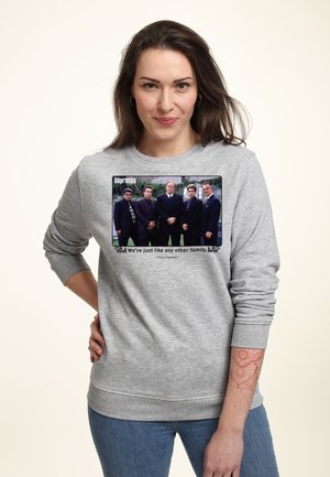 Kobieta w szarej bluzie z nadrukiem zdjęcia obsady Sopranos i cytatem „Jesteśmy jak każda inna rodzina”, stoi z ręką na biodrze.