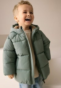 Gepolsterte Kinderjacke in sanftem Grün mit Reißverschluss vorne, Kapuze und gesteppter Textur. Kombiniert mit einem beigen Oberteil und hellblauen Jeans.