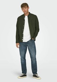 Camisa de botones de manga larga verde oscura sobre una camiseta blanca, combinada con unos vaqueros azules y zapatillas negras. La camisa cuenta con un bolsillo en el pecho.