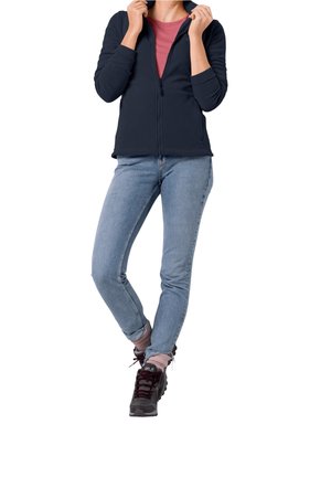 Person iført en navy lynlåstrøje, rød skjorte, lyseblå jeans, pinke sokker og sorte vandresko med det ene ben krydset foran.