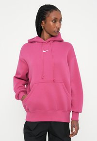 Roza kapuca s vrvicami, kenguru žepom in rebrastimi manšetami. Ima mali beli Nike logo na prsih. Mehka tekstura, oversize kroj.