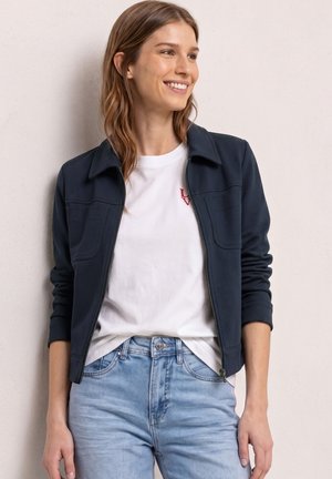 Młoda kobieta uśmiecha się, nosi granatową kurtkę zapinaną na zamek na białą koszulkę i jasnoniebieskie dżinsy, stoi na tle jasnej, gładkiej ściany.