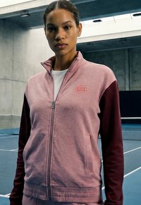 Nő, rózsaszín és bordó cipzáras sportdzsekit viselve, rajta "EAZ" logóval, magabiztosan áll egy fedett teniszpályán.