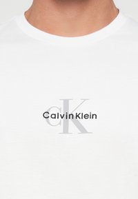 Detailný záber na osobu s bielym tričkom, na ktorom je na hrudi potlačené logo "Calvin Klein" v čiernej a šedej farbe.