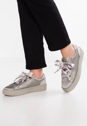 Osoba nosząca metaliczne srebrne niskie trampki z szerokimi satynowymi sznurowadłami, w połączeniu z czarnymi spodenkami do kostek, stojąca na białej podłodze.