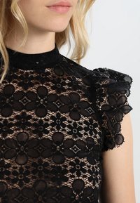 Musta pitsitoppi, jossa on kukkakuviot, korkea kaula-aukko ja lyhyet röyhelöpäät. Materiaali on läpinäkyvä ja hienosti yksityiskohtainen.