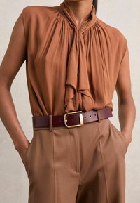 Blusa sin mangas marrón con un cuello anudado drapeado, combinada con pantalones de cintura alta beige y un cinturón marrón oscuro con una hebilla dorada.