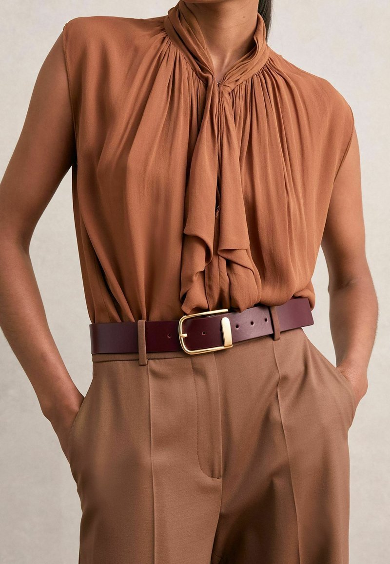 Blusa sin mangas marrón con un cuello anudado drapeado, combinada con pantalones de cintura alta beige y un cinturón marrón oscuro con una hebilla dorada.