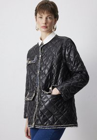 Black quilted jacket with decorative trim, featuring a pointed collar, two front pockets, and a button closure. Smooth, shiny fabric.