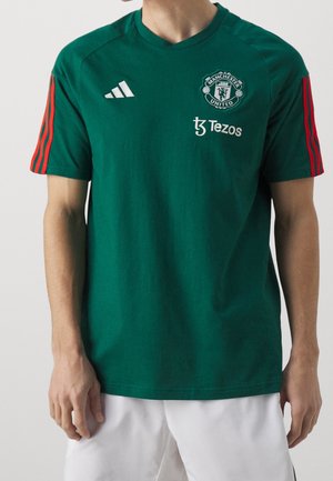 Personne portant un maillot vert de Manchester United avec des rayures rouges sur les manches, des logos blancs et un short blanc, vue des épaules aux cuisses.