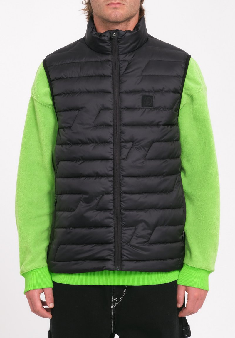 Gilet imbottito nero con colletto alto, caratterizzato da dettagli di cuciture geometriche, indossato sopra una maglietta a maniche lunghe di colore verde brillante.