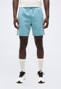 Άνδρας που φοράει ανοιχτό μπλε σορτς με κορδόνι, λευκές κάλτσες και λευκά αθλητικά, στέκεται μπροστά σε απλό φωτεινό φόντο.