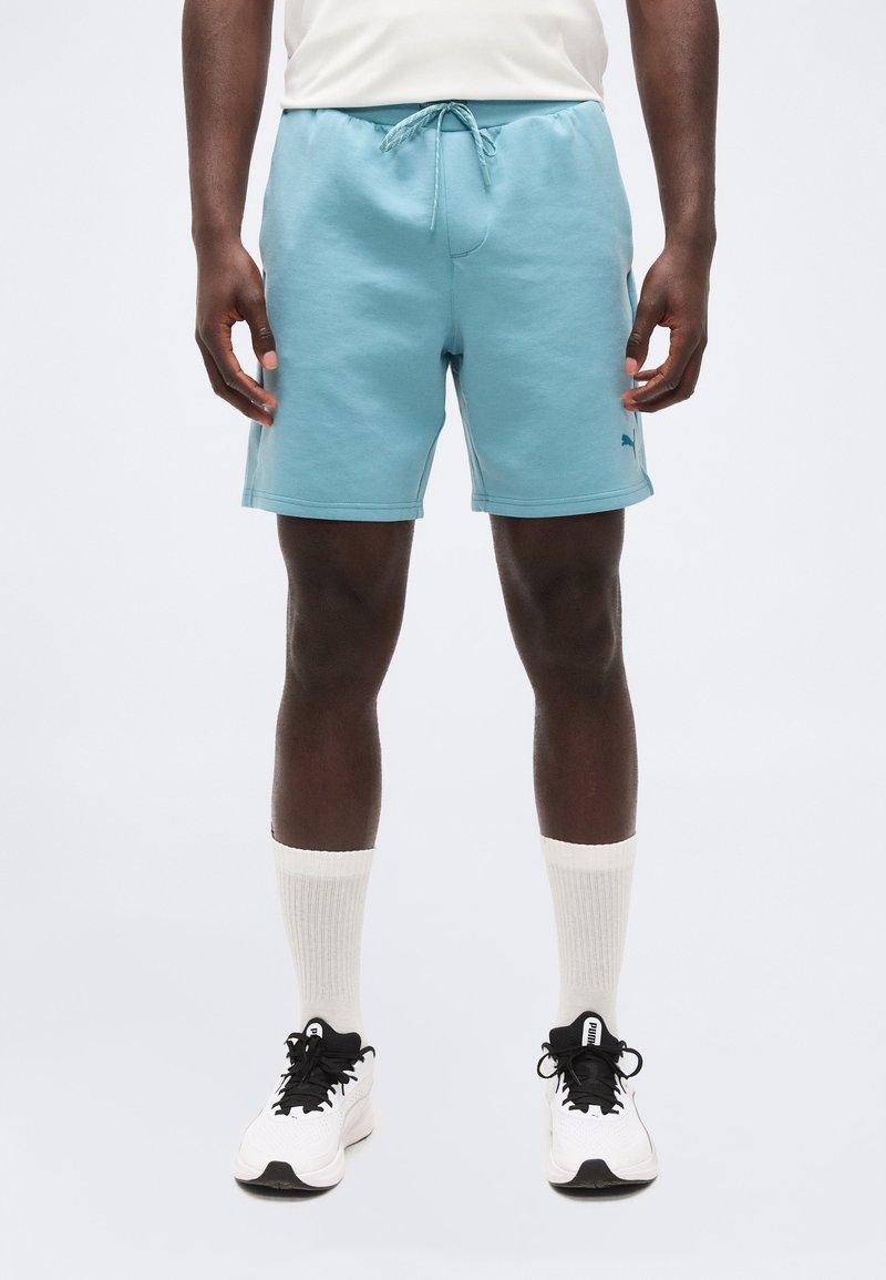 Άνδρας που φοράει ανοιχτό μπλε σορτς με κορδόνι, λευκές κάλτσες και λευκά αθλητικά, στέκεται μπροστά σε απλό φωτεινό φόντο.