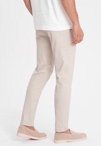 Mężczyzna w beżowych dopasowanych spodniach i pasujących zamszowych mokasynach, stojący z lekko ugiętą nogą na białym tle.