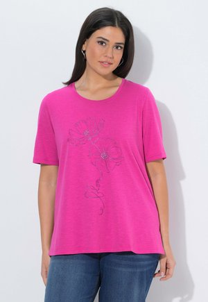 Kobieta o ciemnych włosach, nosząca kolczyki koła, jasnoróżową koszulkę z krótkim rękawem zdobioną wzorem kwiatowym oraz niebieskie dżinsy, stojąca na tle białej ściany.