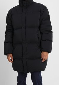 Giubbotto imbottito nero con collo alto, sezioni trapuntate per l'isolamento, tasche laterali con cerniera e dettaglio logo sul petto.