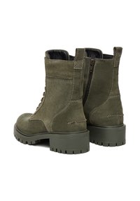 Olīvu zaļi zamša šņorējamie zābaki ar sānu rāvējslēdzējiem un masīvām gumijas zolēm, skatīti no aizmugures un slīpi.