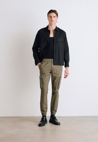 Camisa negra de botones, camiseta negra ajustada, pantalones cargo verde olivo con bolsillos laterales y botas negras con cordones. Diseño simple y moderno.