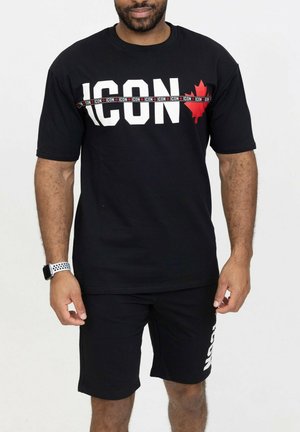 Homme portant un t-shirt noir avec l'inscription « ICON » et une feuille d'érable rouge, assorti d'un short noir avec un texte blanc sur la cuisse gauche.