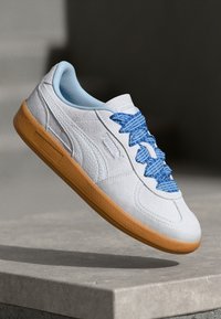 Zapatilla de piel blanca con un cuello azul claro, que presenta cordones de patrón azul y una suela de goma. Diseño simple con detalles texturizados.