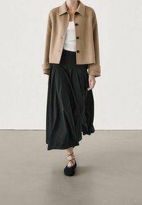 Brown cropped jacket with black buttons, white top, and flowing black maxi skirt. Model wears black lace-up flats. Smooth textures and layered look.