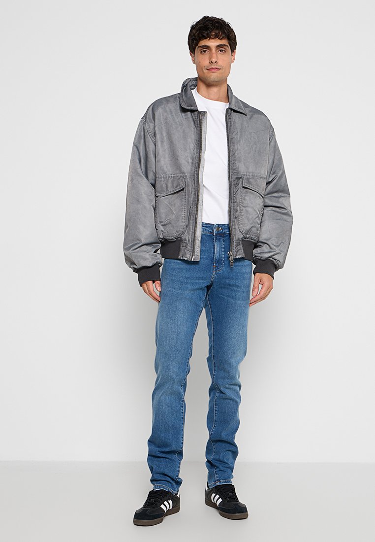 Szara kurtka bomberka o gładkiej teksturze, zapinana na zamek, z dwiema przednimi kieszeniami, zestawiona z niebieskimi dżinsami i czarnymi trampkami.