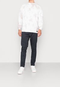 Chemise blanche à manches longues avec un léger motif tie-dye rose, associée à un pantalon noir slim et des baskets blanches avec des accents roses.