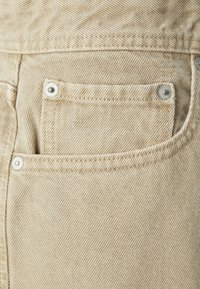 Tela de mezclilla beige con textura diagonal, que presenta un pequeño bolsillo y detalles de botones metálicos. Bordes cosidos visibles para mayor durabilidad.